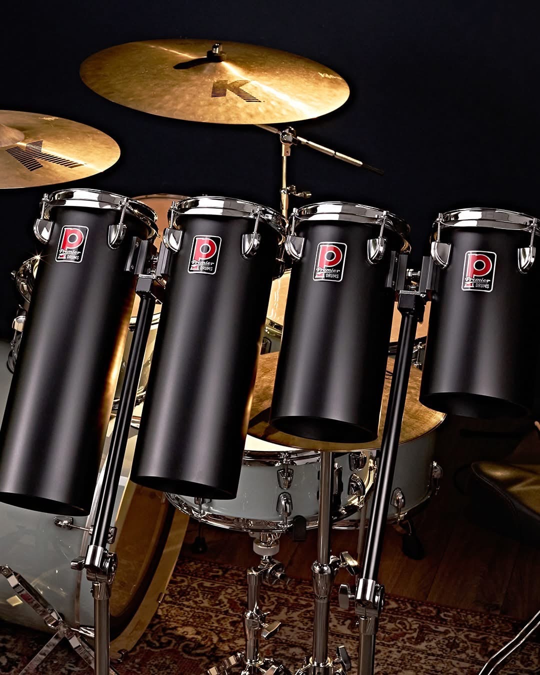 Four Premier Artist Octobans in matte black with chrome triple-flange hoops and red Premier badges, mounted in a row in front of a drum kit