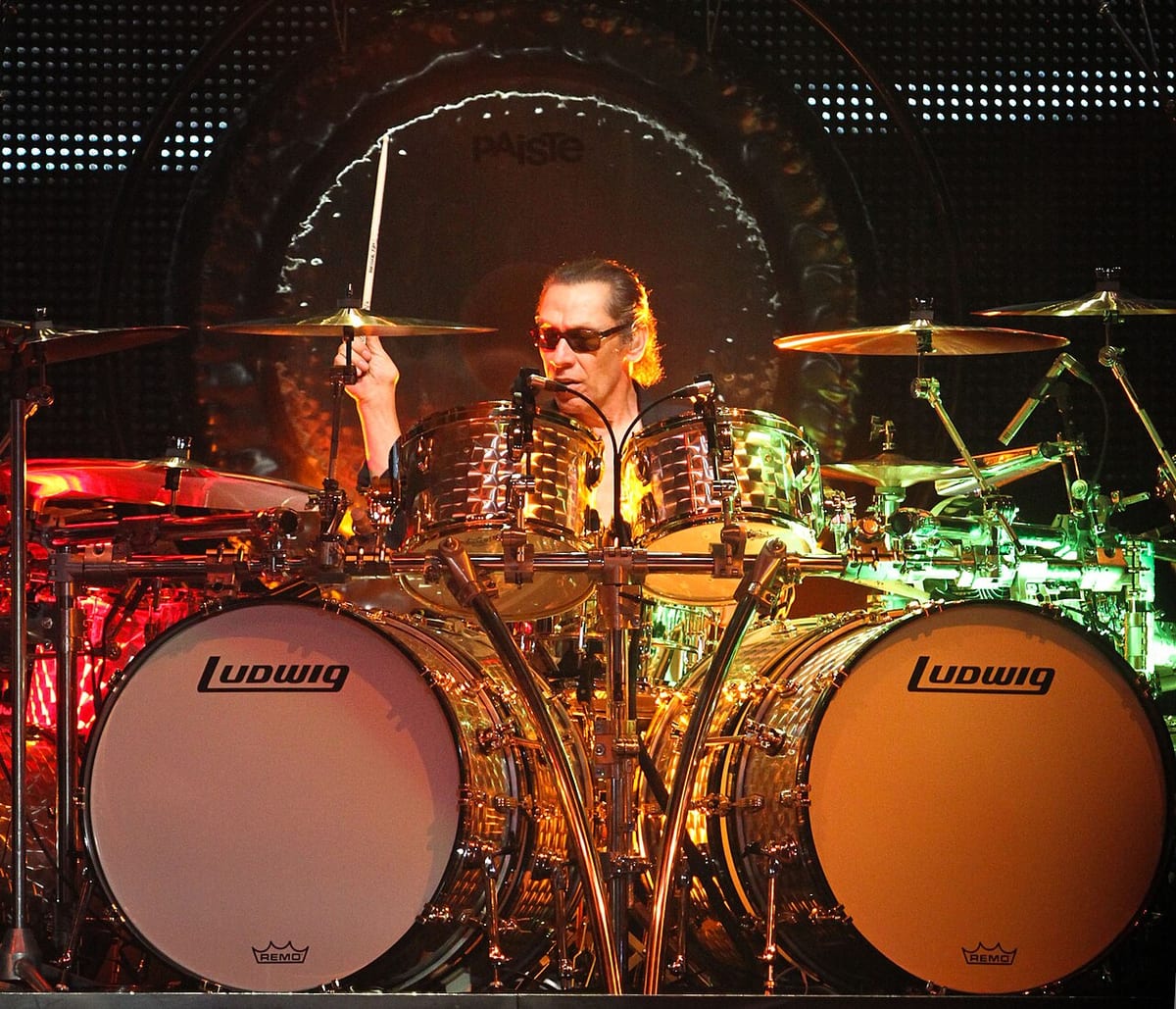 Alex Van Halen in sunglasses behind twin Ludwig bass drums and a brass kit, with a large Paiste gong behind him