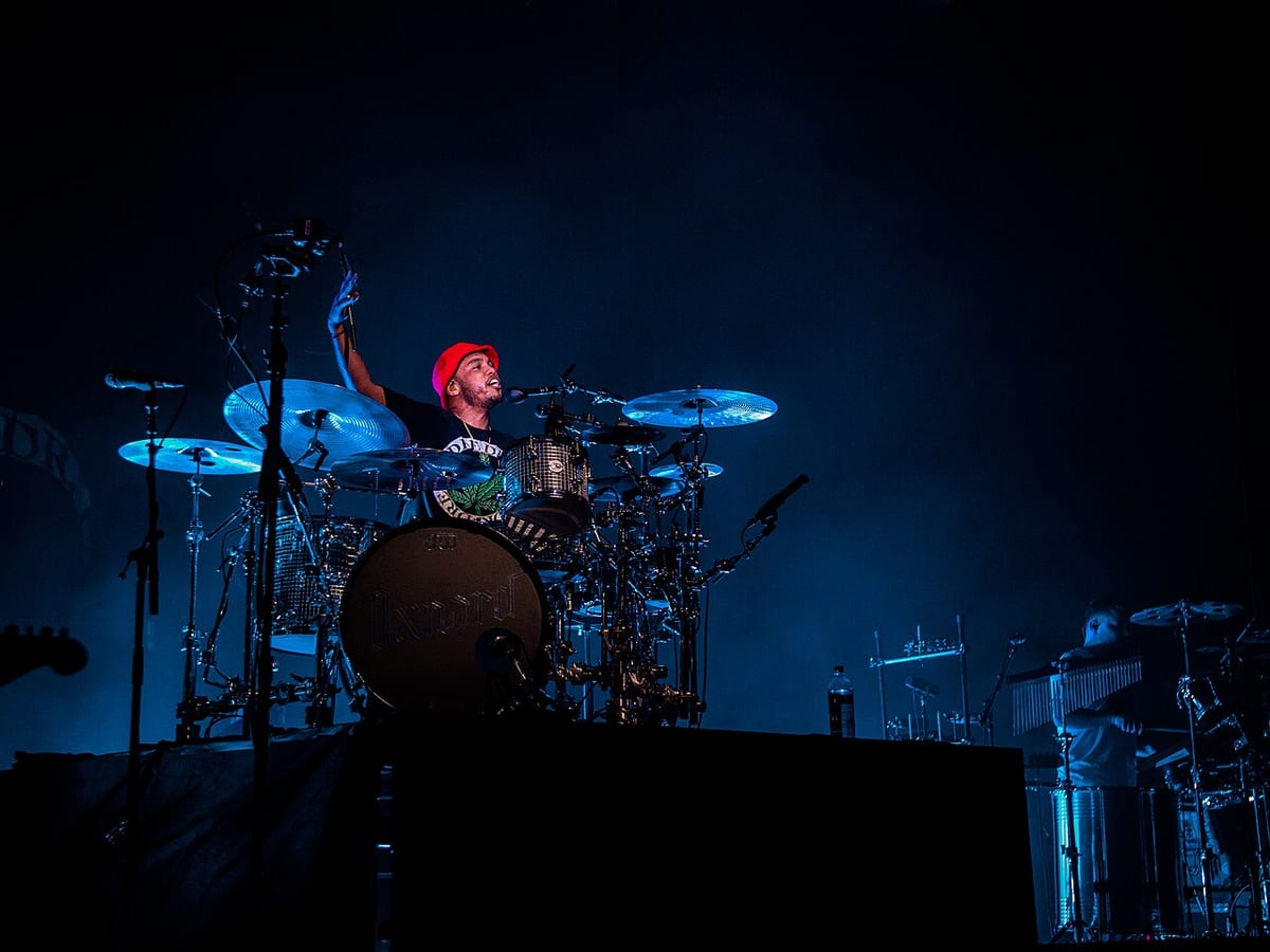 Anderson .Paak in a red beanie, singing into a microphone while playing a kit with Oxnard written across the bass drum head