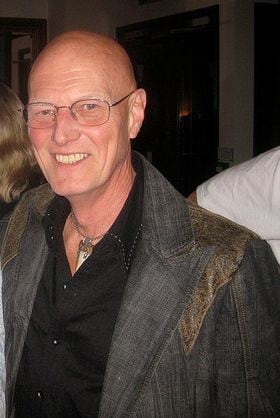 Chris Slade, bald and smiling in glasses and a grey jacket, photographed at an event