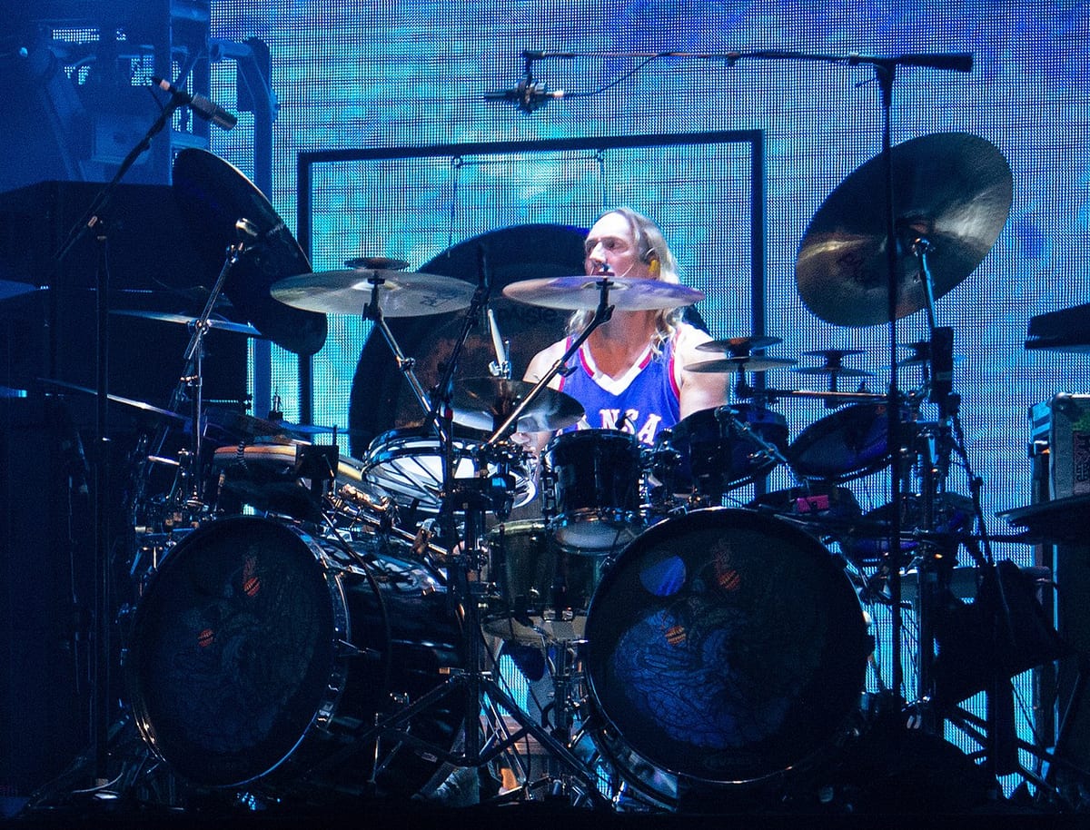 Danny Carey seated behind his Sonor kit under blue stage light, surrounded by Paiste cymbals and twin bass drums