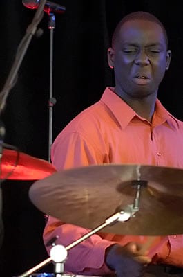 Larnell Lewis in a salmon shirt, eyes down, playing behind the ride cymbal