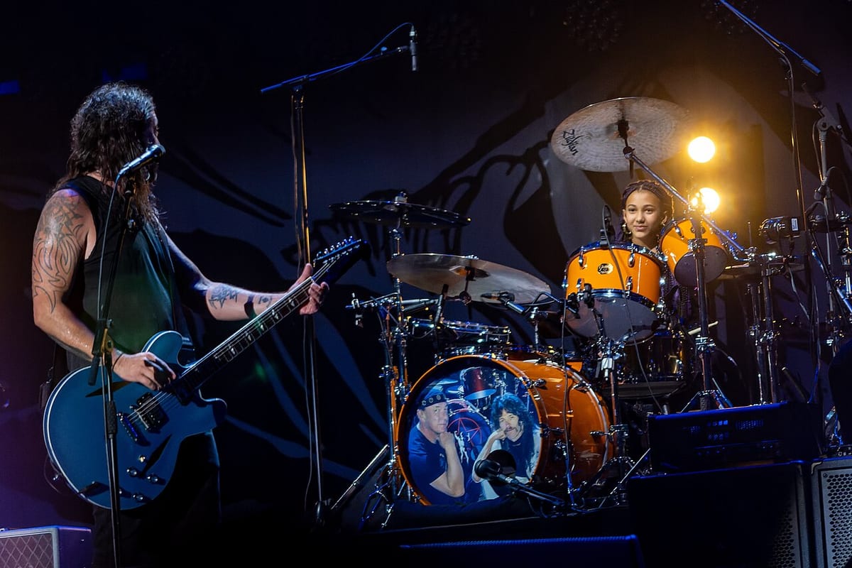 Nandi Bushell behind an orange kit on a stadium stage, smiling across at Dave Grohl on guitar
