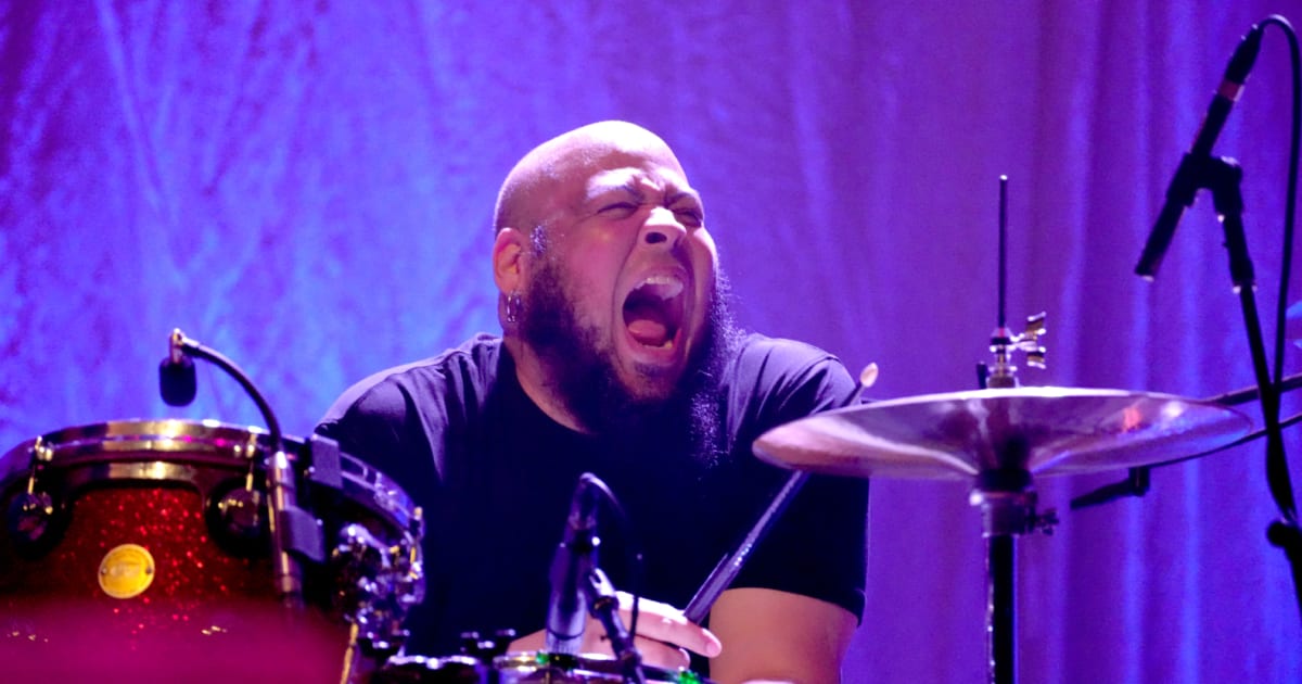 A drummer mid-shout behind a red sparkle kit under purple stage lights