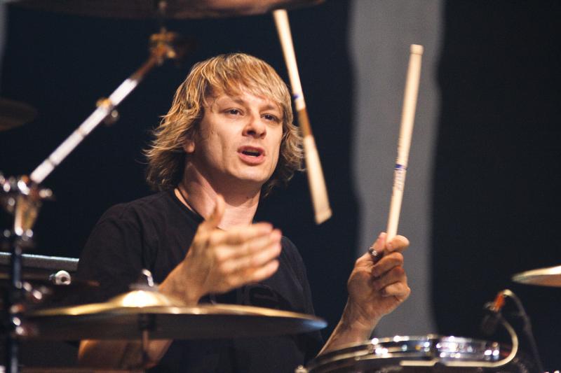 Ray Luzier behind his kit in a black T-shirt, one stick raised mid-song, surrounded by cymbals
