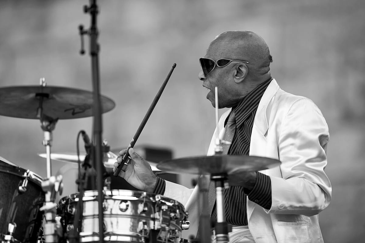 Roy Haynes in a white suit and sunglasses, stick raised over the ride cymbal, playing outdoors