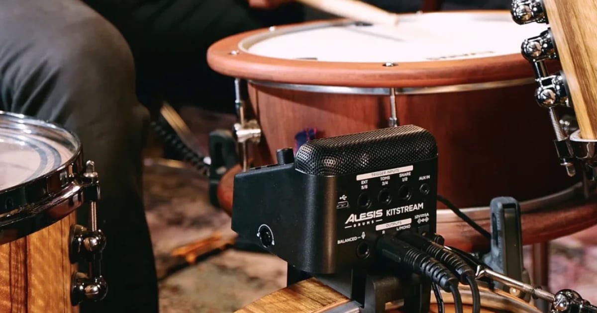 The black Alesis KitStream unit clamped to a wooden bass drum hoop, with cables in its trigger inputs and outputs and a drummer playing a snare behind it