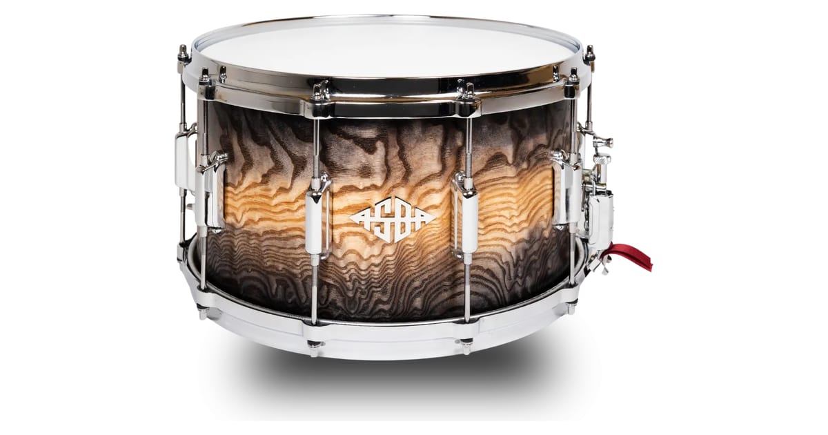The ASBA Signature Rémi Serafino 14x8 snare, with a Tamo ash shell in a dark-to-natural burst, chrome tube lugs and the diamond ASBA badge